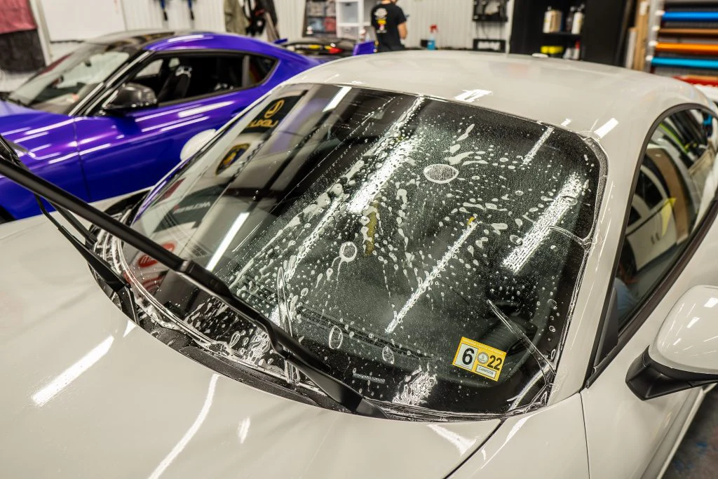 A car in a workshop is undergoing a windshield cleaning process with soap and water bubbles on the glass. The windshield wipers are raised, and another car, possibly getting Paint Protection Film applied, is visible in the background.