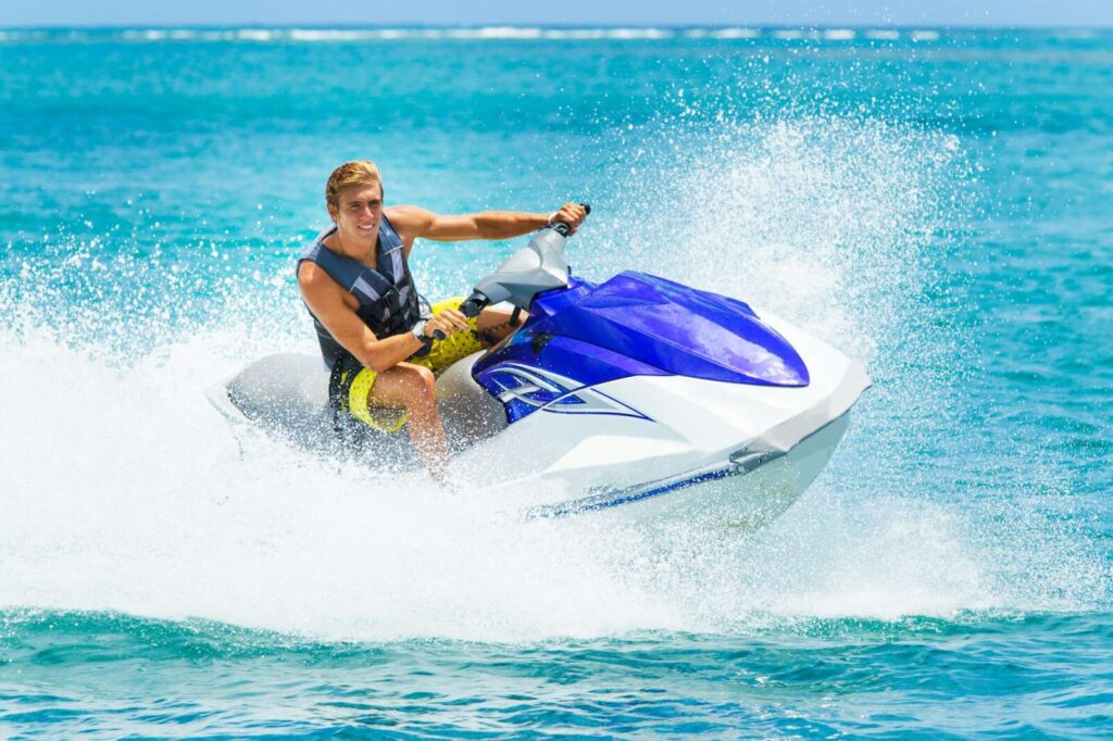 A person riding a blue and white jet ski, protected by paint protection film, creates a splash in the ocean, wearing a black life vest and yellow shorts.