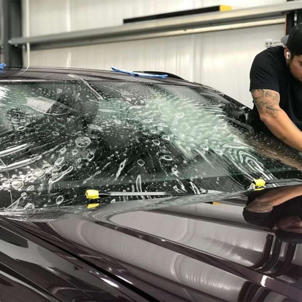 A person applies a Paint Protection Film to a car's windshield, attaching it carefully to ensure smooth placement.