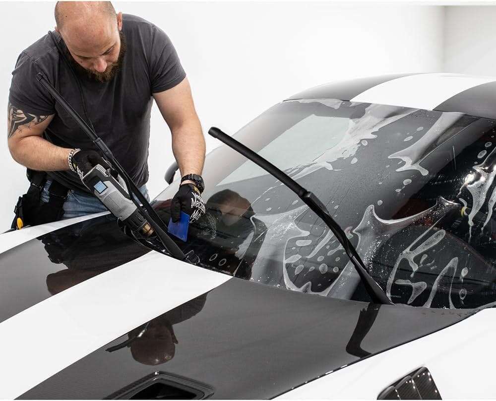 In a bustling workshop, an expert meticulously applies paint protection film to a car's hood and windshield, ensuring every curve is shielded with precision.